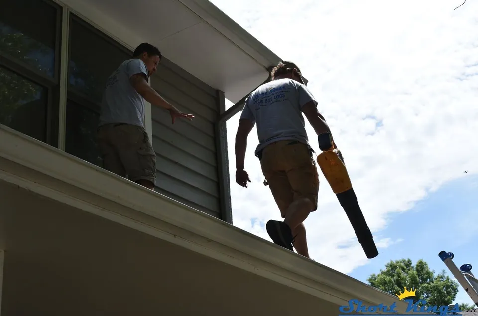 Two people on a lower roof, one carrying a yellow blower