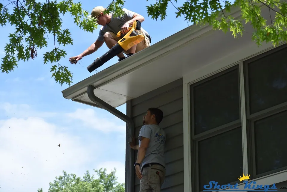 Two people working on gutters, one on the roof and one below at the wall